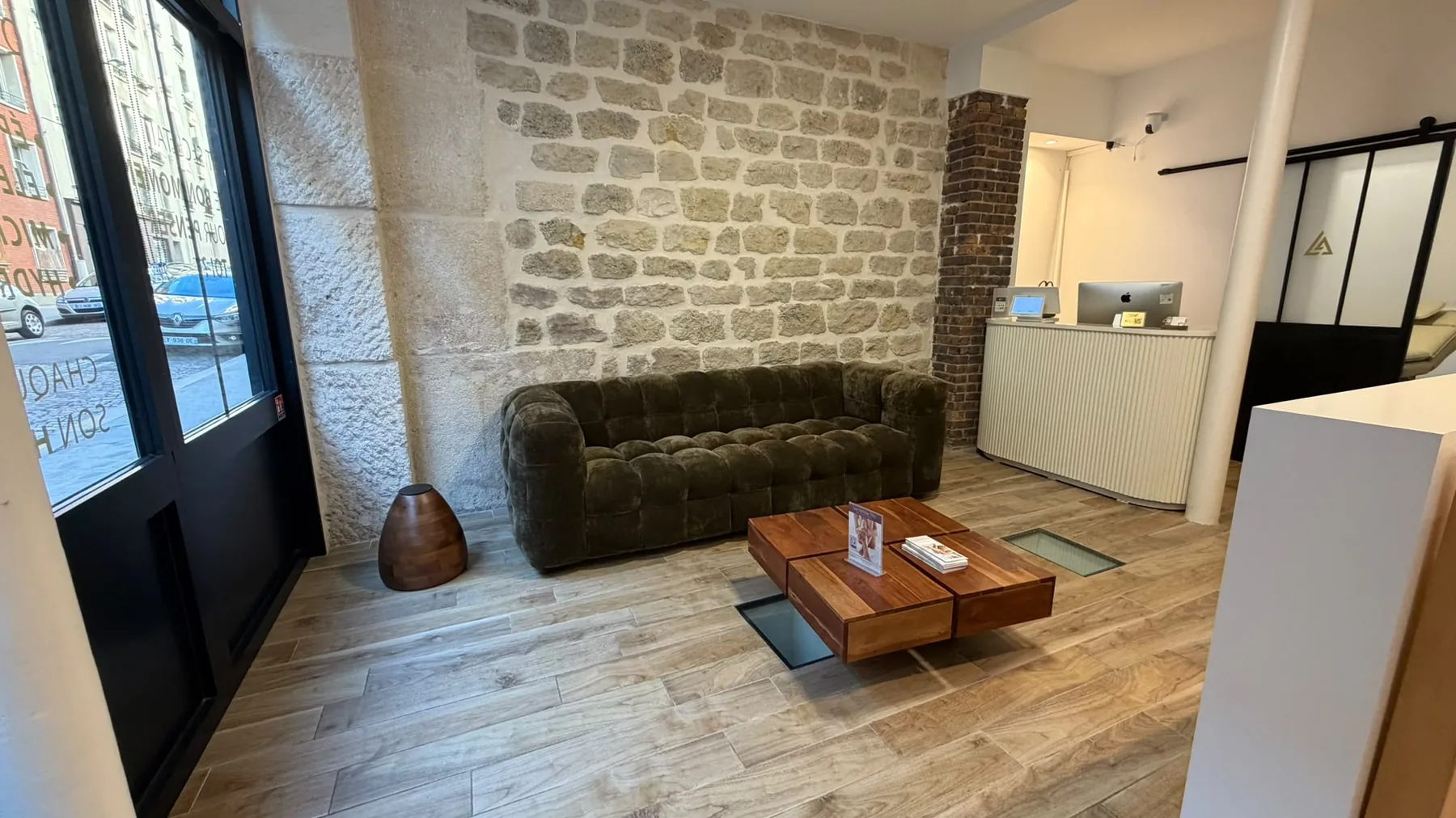 L'accueil de l'Atelier Laser 17 : mur de pierre apparente, canapé en velours vert, table basse en bois et comptoir d'accueil